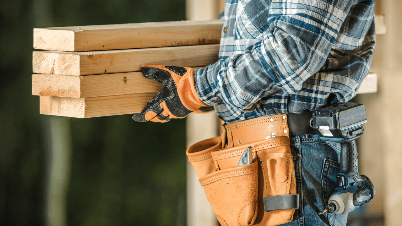 Carpenter wearing orange work gloves carrying a stack of 2x4s.